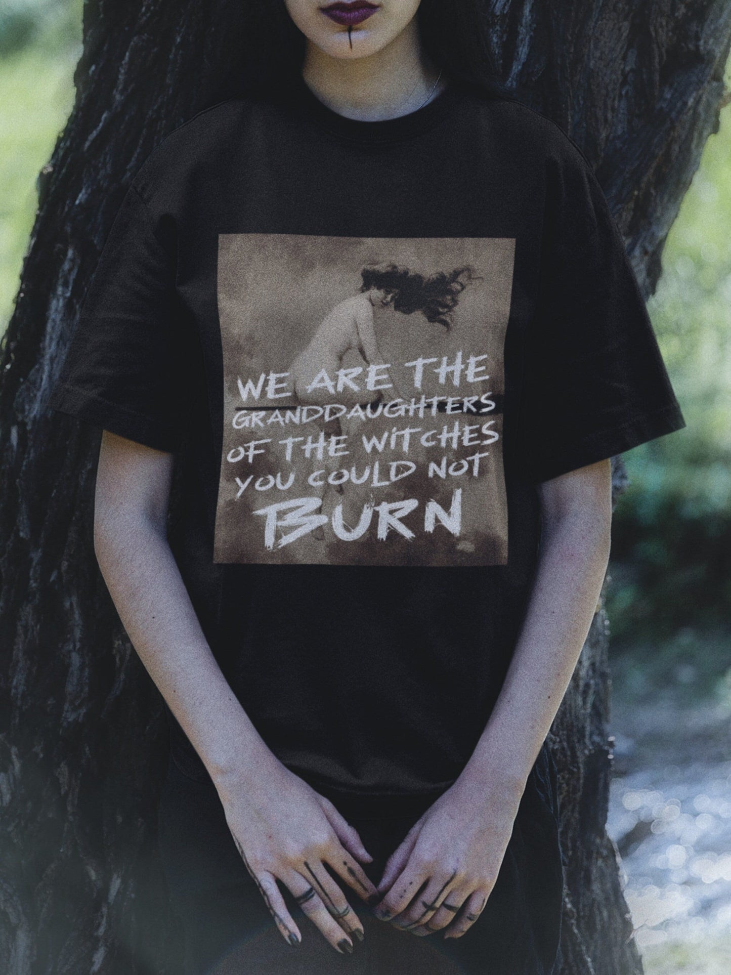 a goth wiccan woman wears an oversized black tshirt with the image of a witch riding a broom and the words "we are the granddaughters of the witches you could not burn" in a brush font over the image