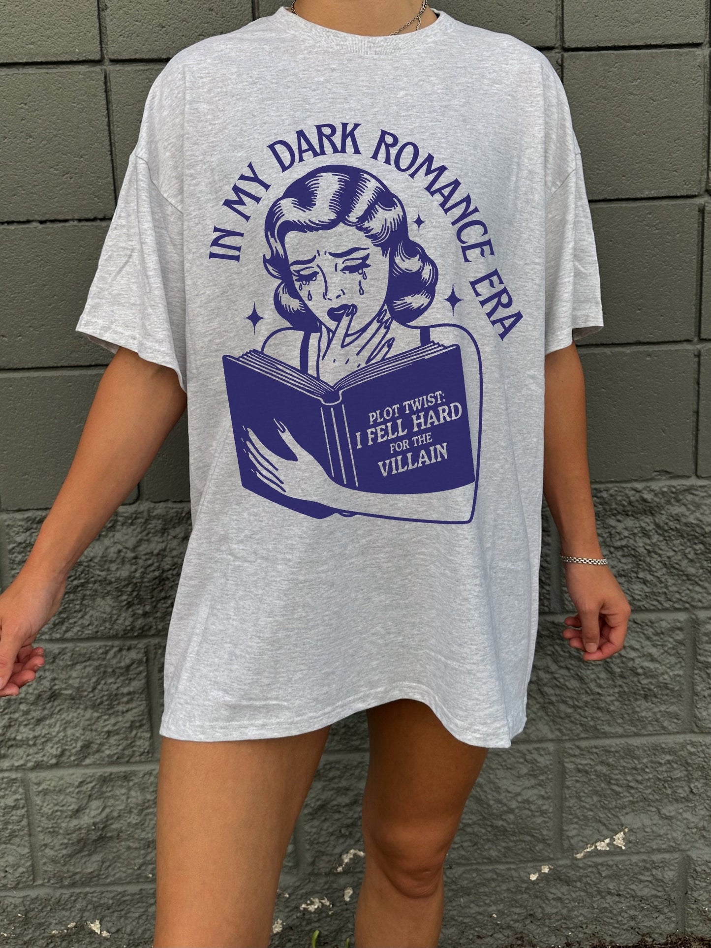 May include: A gray t-shirt with a blue graphic of a woman reading a book. The book has the text 'Plot Twist: I Fell Hard For The Villain' on it. The text 'In My Dark Romance Era' is written around the graphic.