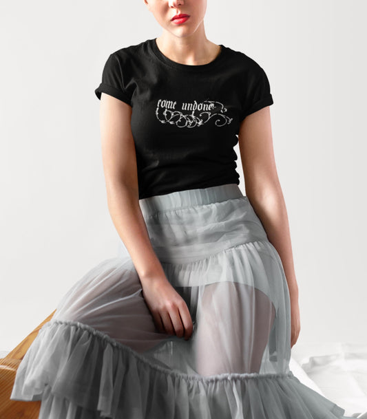 May include: Black t-shirt with the words "come undone" in white script. The shirt is paired with a light gray, layered tulle skirt. The model is seated on a wooden surface.
