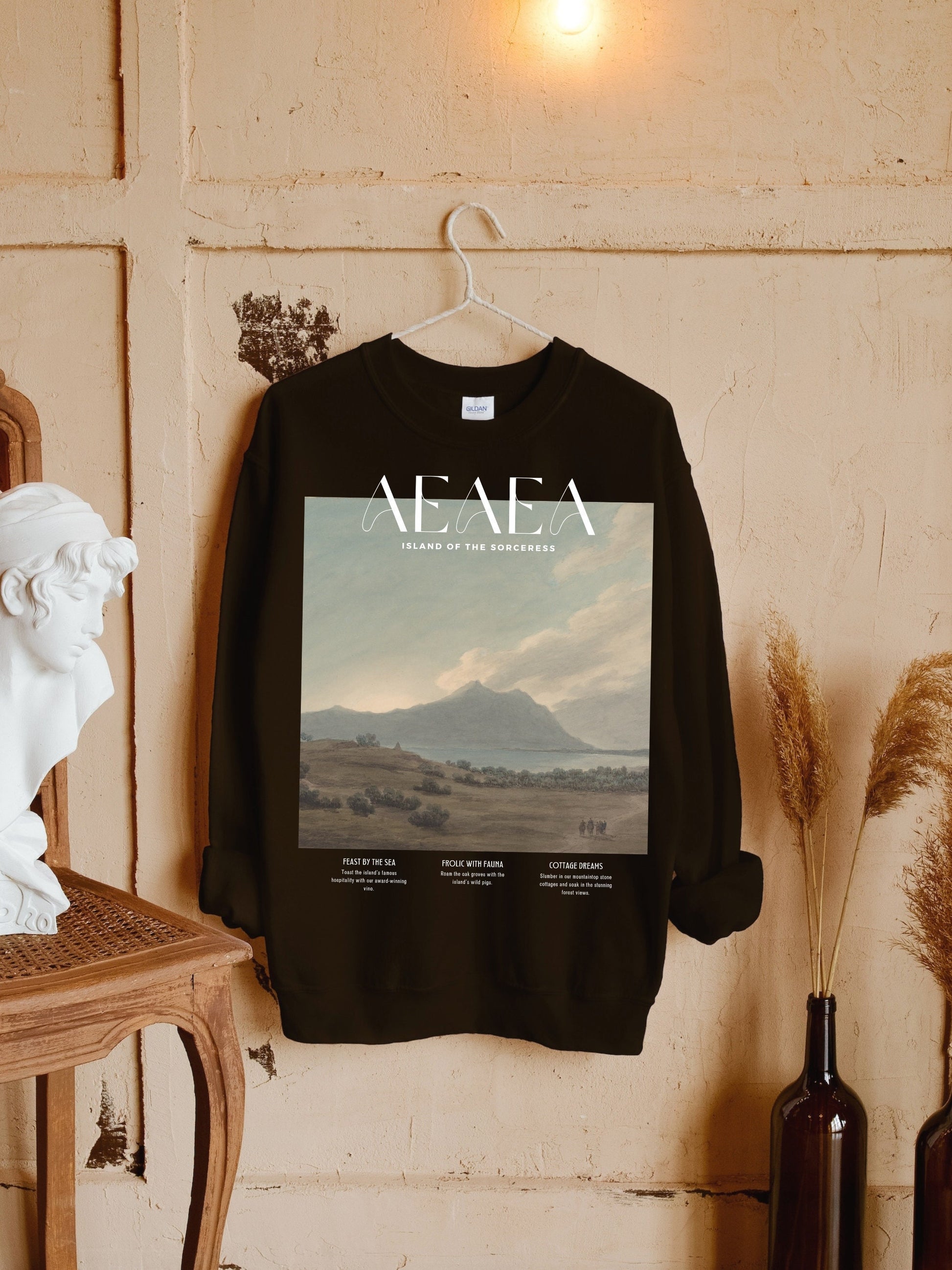 May include: Black sweatshirt with a graphic print of a landscape scene with mountains, water, and a group of people walking. The text "Aeaea Island of the Sorceress" is printed above the image. The text "Feast by the Sea", "People with Fauna", and "Cottage Dreams" is printed below the image.