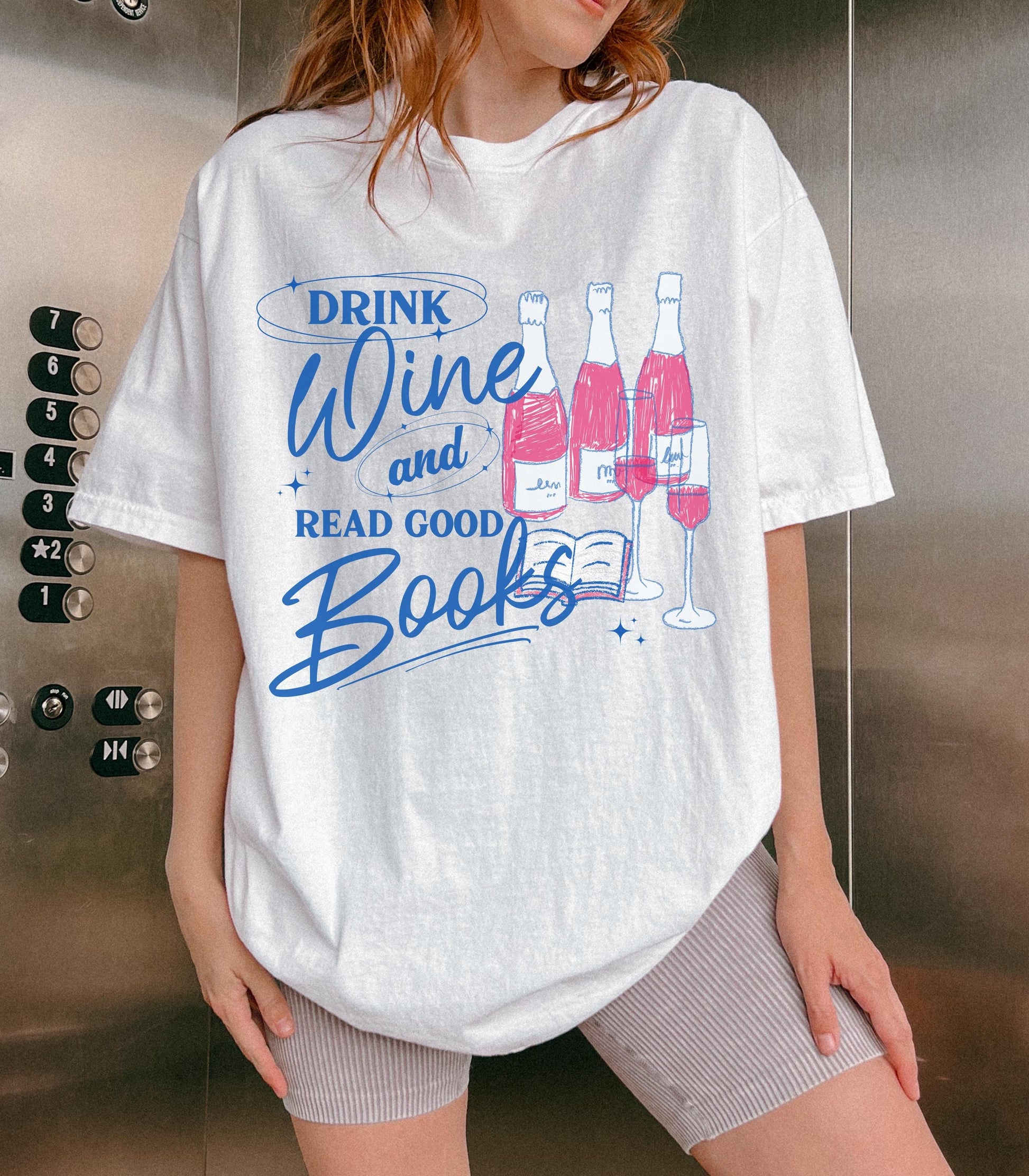 May include: White t-shirt with a blue and pink graphic. The graphic features the words "DRINK Wine and READ GOOD Books" with illustrations of wine bottles, a wine glass, and an open book. The shirt is a casual, oversized fit.