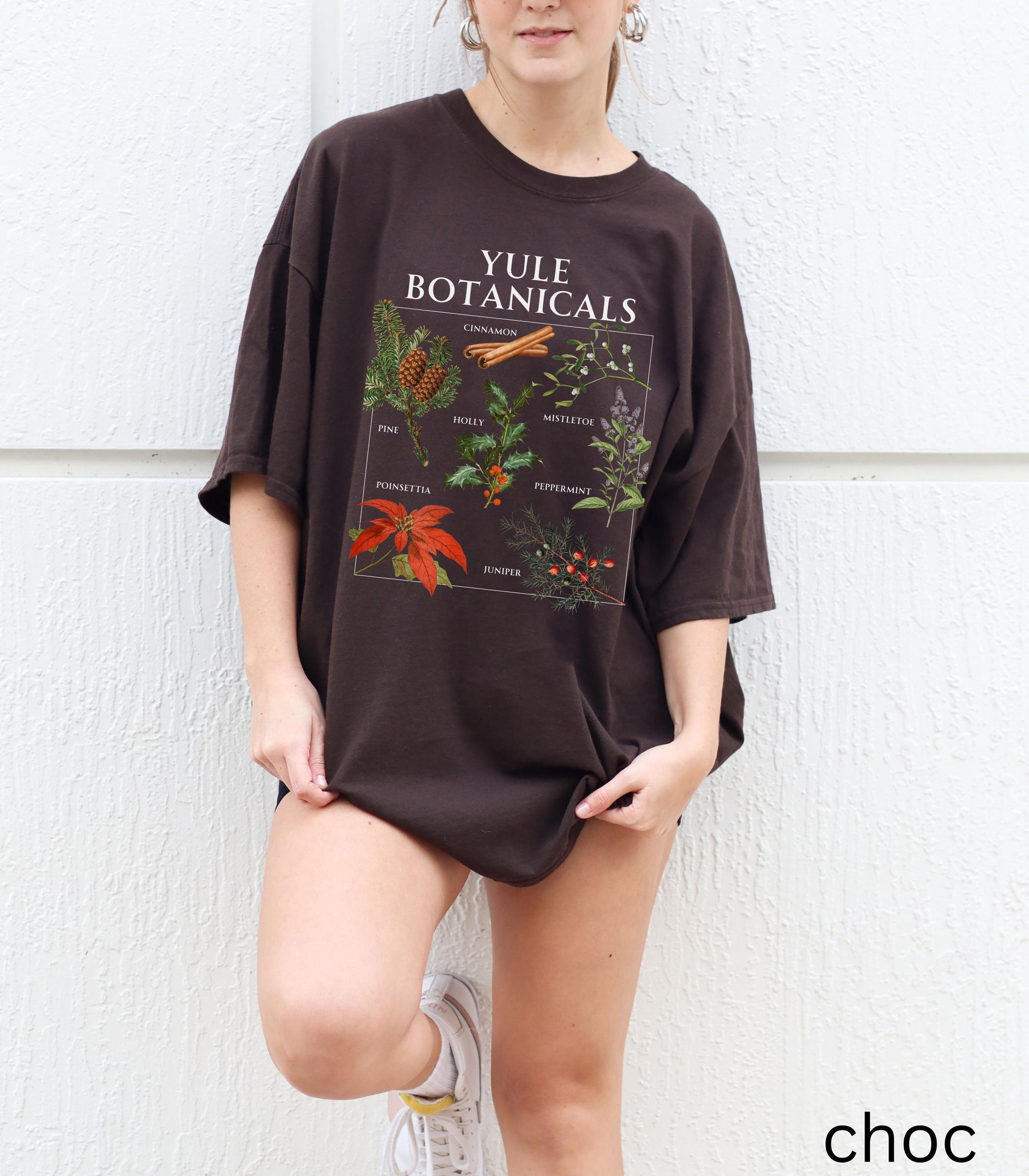 May include: A dark brown t-shirt with the text "YULE BOTANICALS" and botanical illustrations. The shirt features images of pine, cinnamon, holly, mistletoe, poinsettia, peppermint, and juniper.