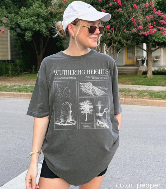 May include: Pepper-colored t-shirt with the text "Wuthering Heights" and illustrations from the novel. The shirt features a crew neck and short sleeves. The model is wearing a white baseball cap and sunglasses.